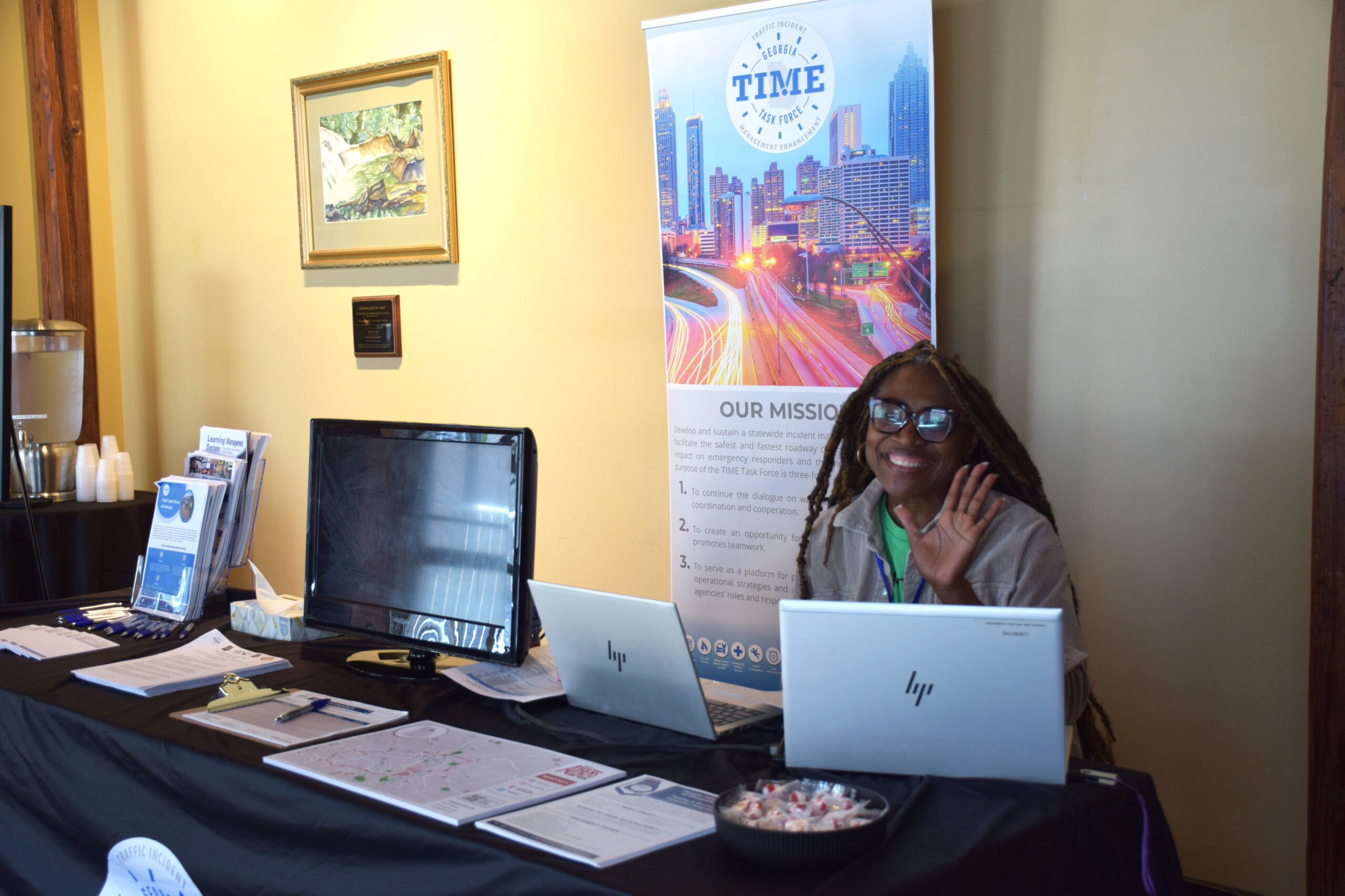 The TIME Task Force Booth at an event with promotional material; the woman working the table smiles and waves at the camera. 
