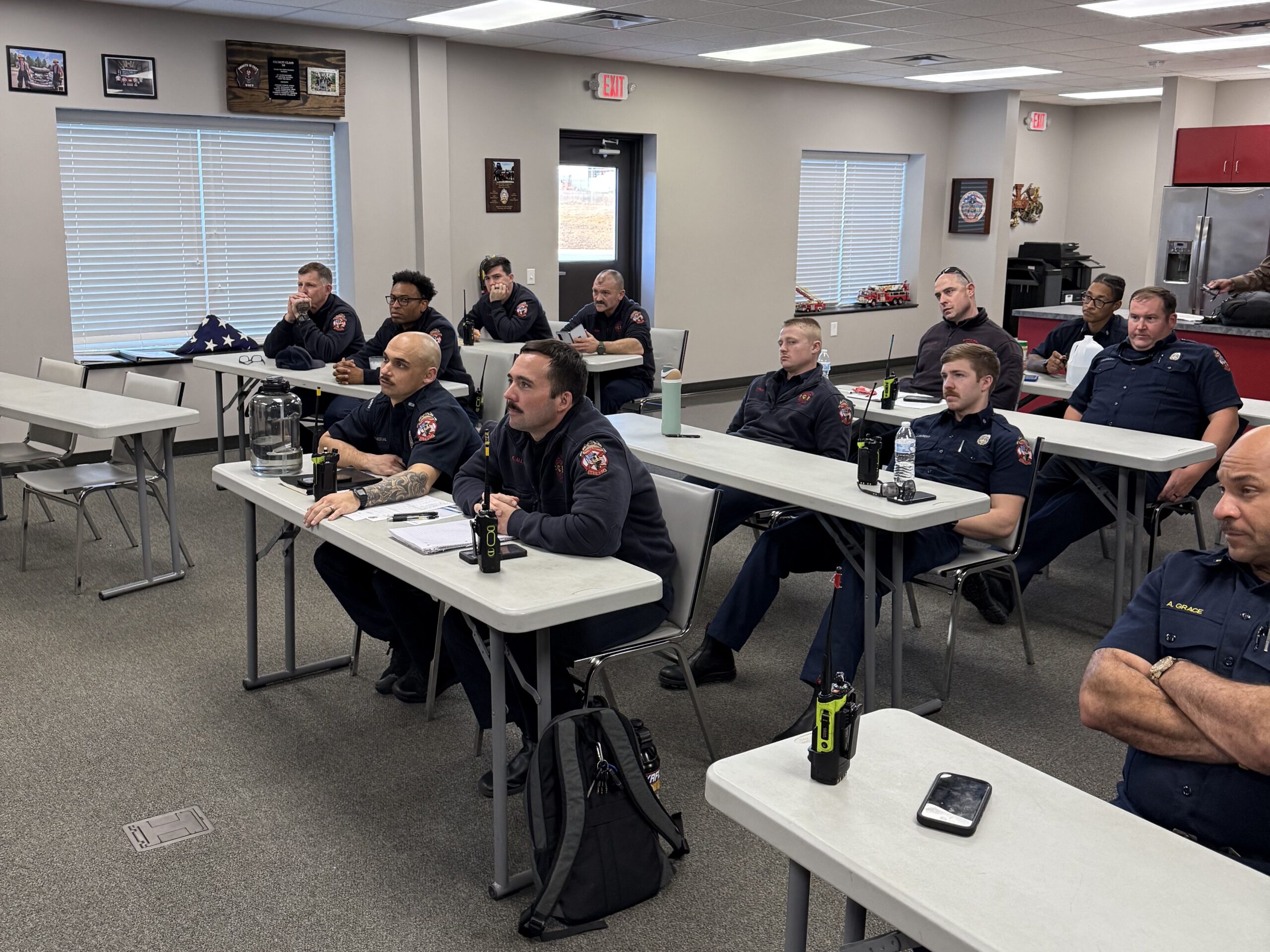 Firefighters sit in a room at multiple tables with their focus on the front of the room, presumably on the presenter and lesson material. 