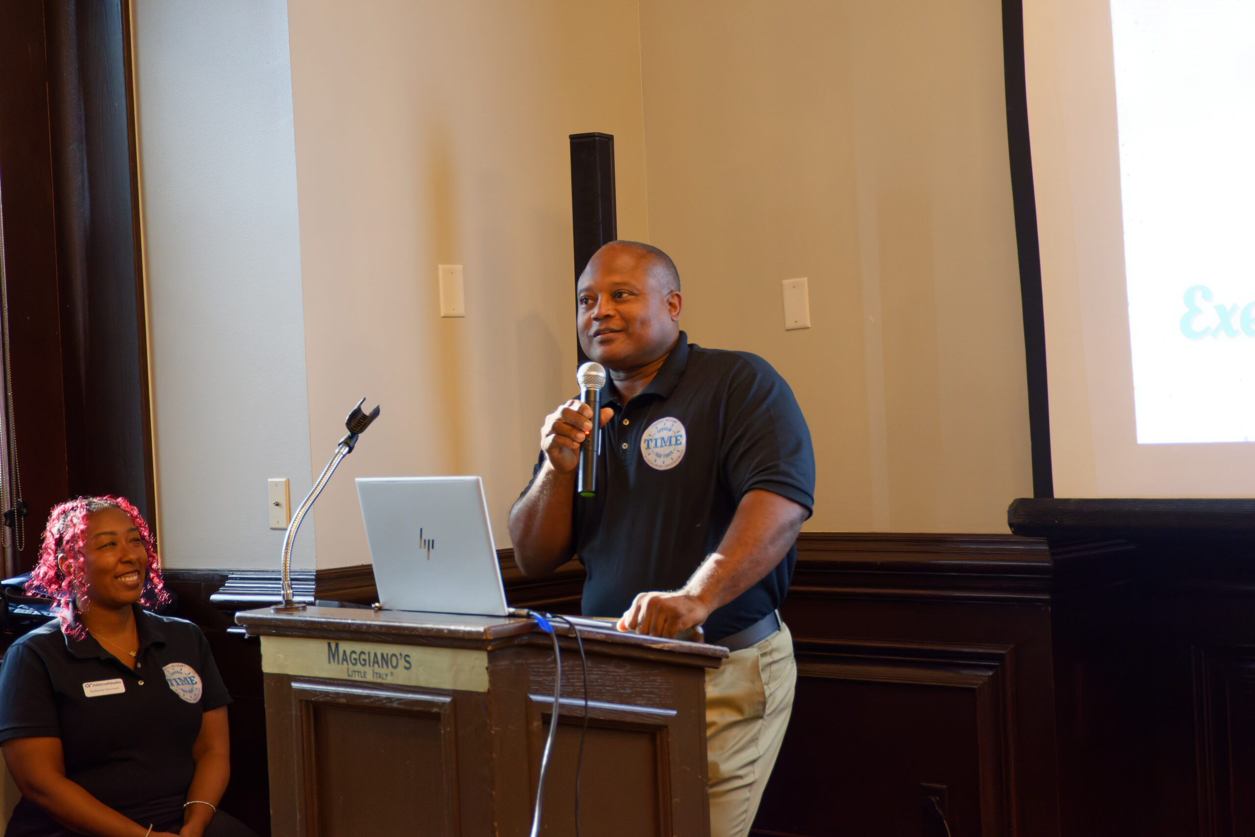 The TIME Task Force co-chair speaks to a non-visible crowd at a podium with a microphone. A TIME Task Force member is sitting and smiling up at the speaker.