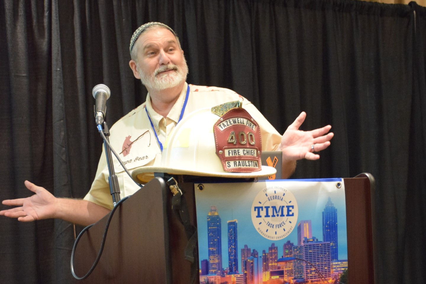 A close up of a speaker from the 2024 Conference. He is standing at a podium with his firefighter helmet.