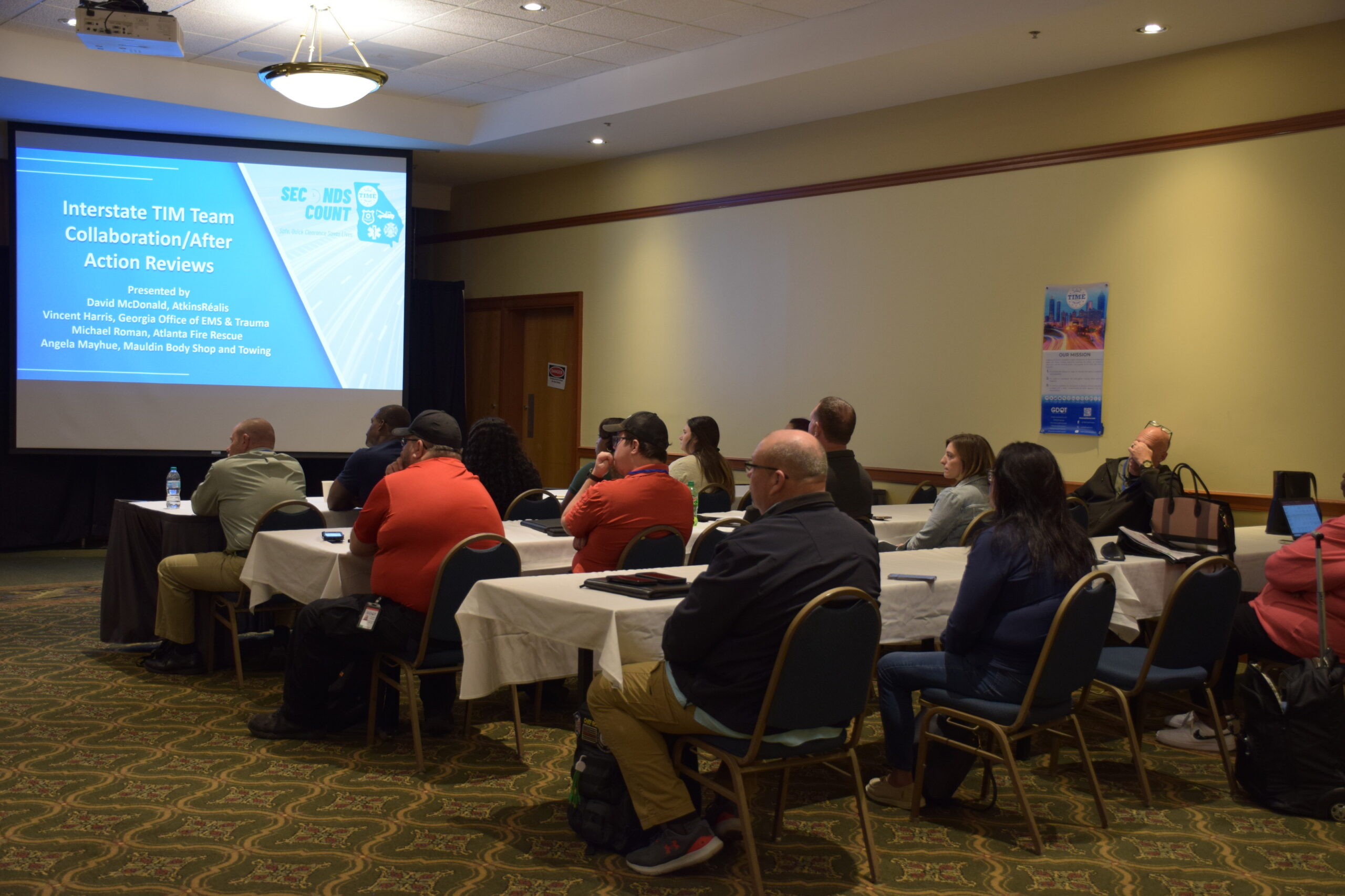 A session from the2024 Conference. Speakers are seated in a full room, looking at a screen with the presentation title.