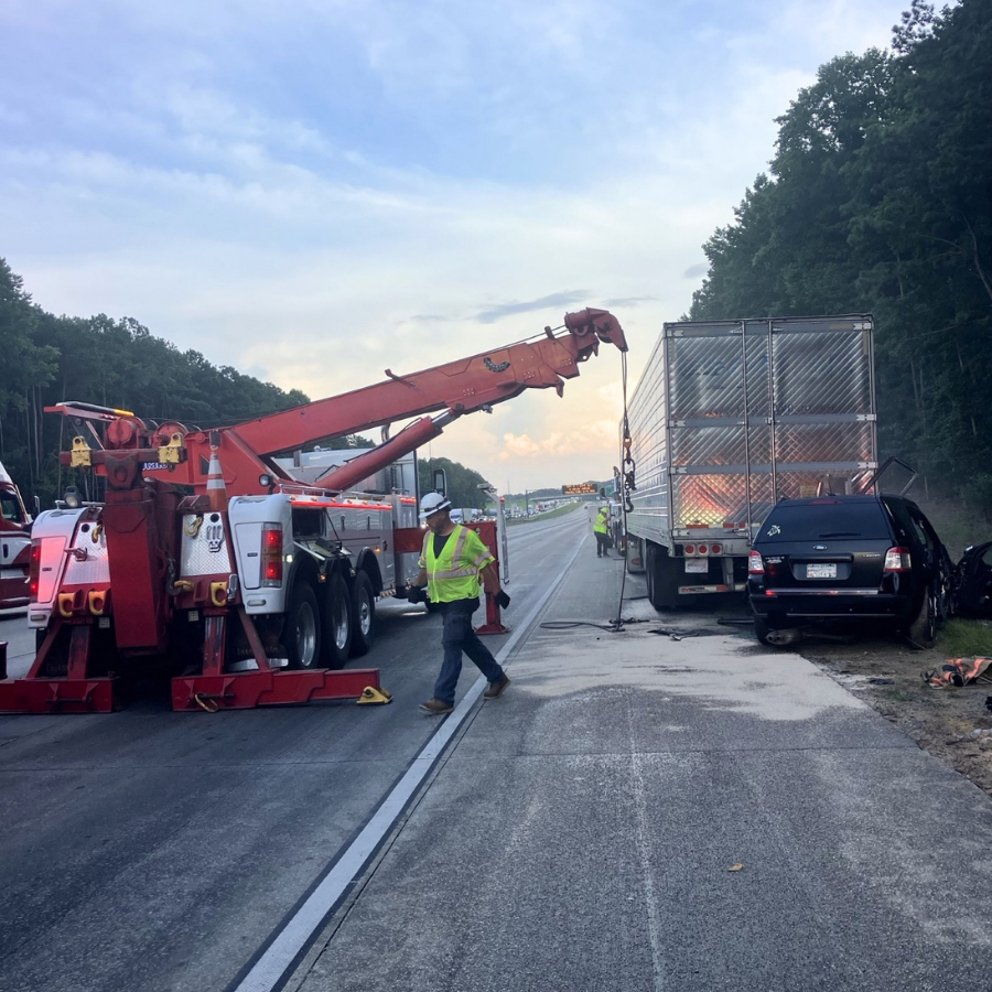 A responder walks behind an incident on the road where a red tow truck is picking up a large semi-truck. A black SUV is crashed into the back of the semi-truck.